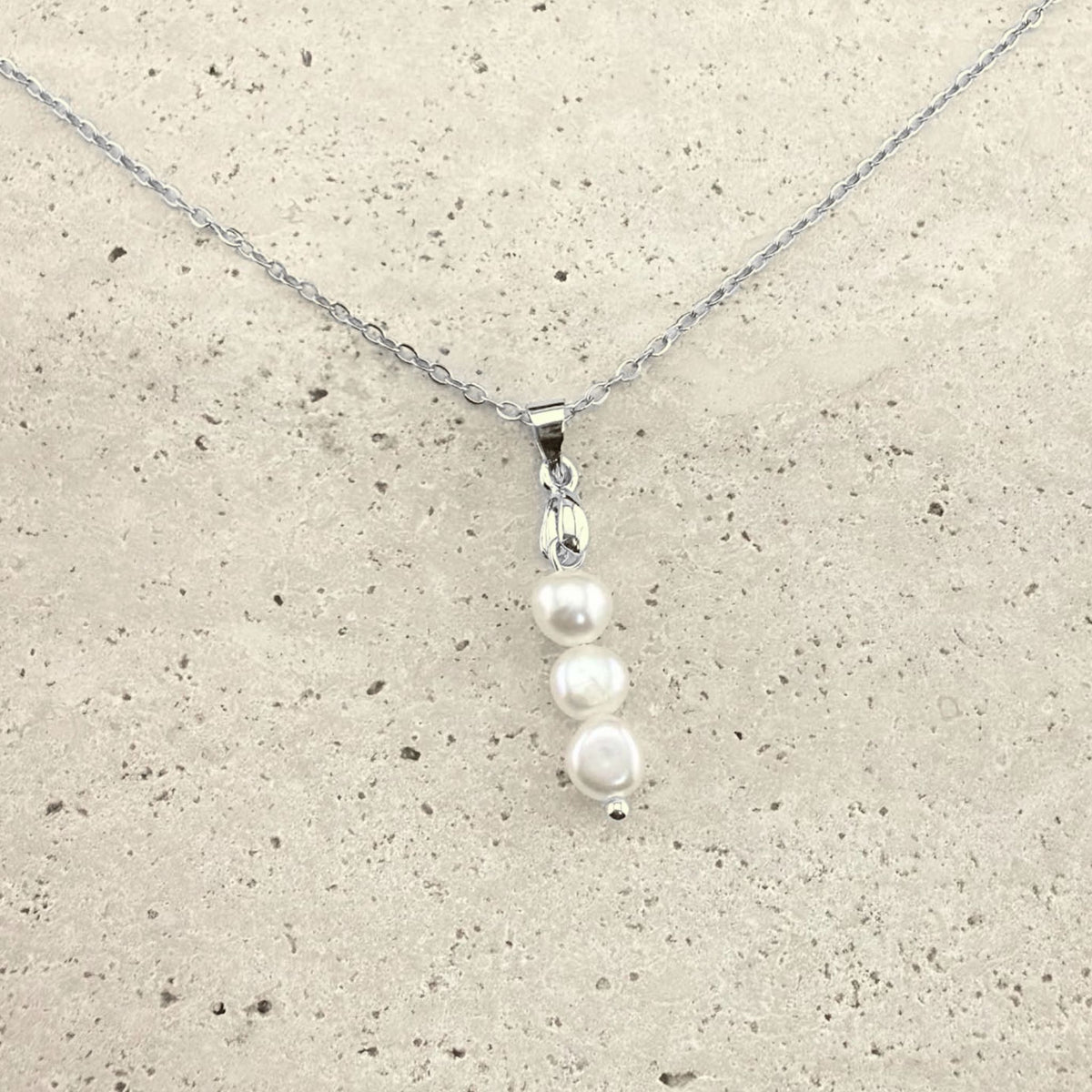 A silver Aking Sinta necklace featuring a delicate pendant composed of three white, organic-shaped baroque pearls stacked vertically. The pearls are suspended from a polished silver bail and a fine silver cable chain, presented on a light, textured natural stone surface