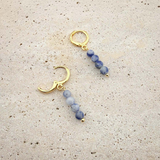 Macro view of a pair of gold Sodalite huggie earrings on marble, with one hinge clasp shown securely open.