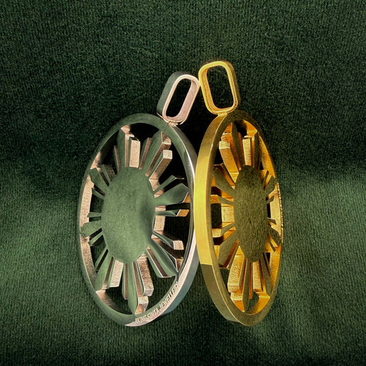 A striking pair of Aking Sinta statement Araw medallions, one in polished silver and one in rich gold, featuring a sun-inspired, filigree-cut design. The medallions are elegantly positioned against a deep, dark green velvet background, highlighting their intricate craftsmanship and reflective metallic finish