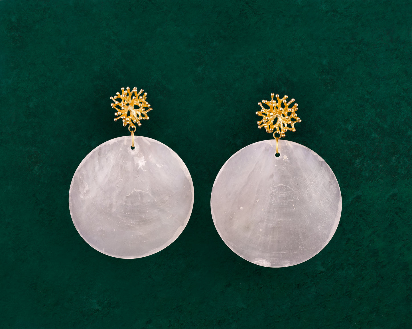 A detailed close-up of a pair of Aking Sinta Statement Capiz and Coral Stud earrings on a dark green background; featuring large, round white Capiz shell disks with a small, round, bright red coral stone fixed at the center of each.