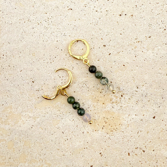 A pair of Aking Sinta gold huggie earrings with three-bead Moss Agate charms on a white textured stone; one gold clasp is open to show the hinge mechanism, while the other is securely closed, showcasing the dark green mossy patterns inside the clear beads.