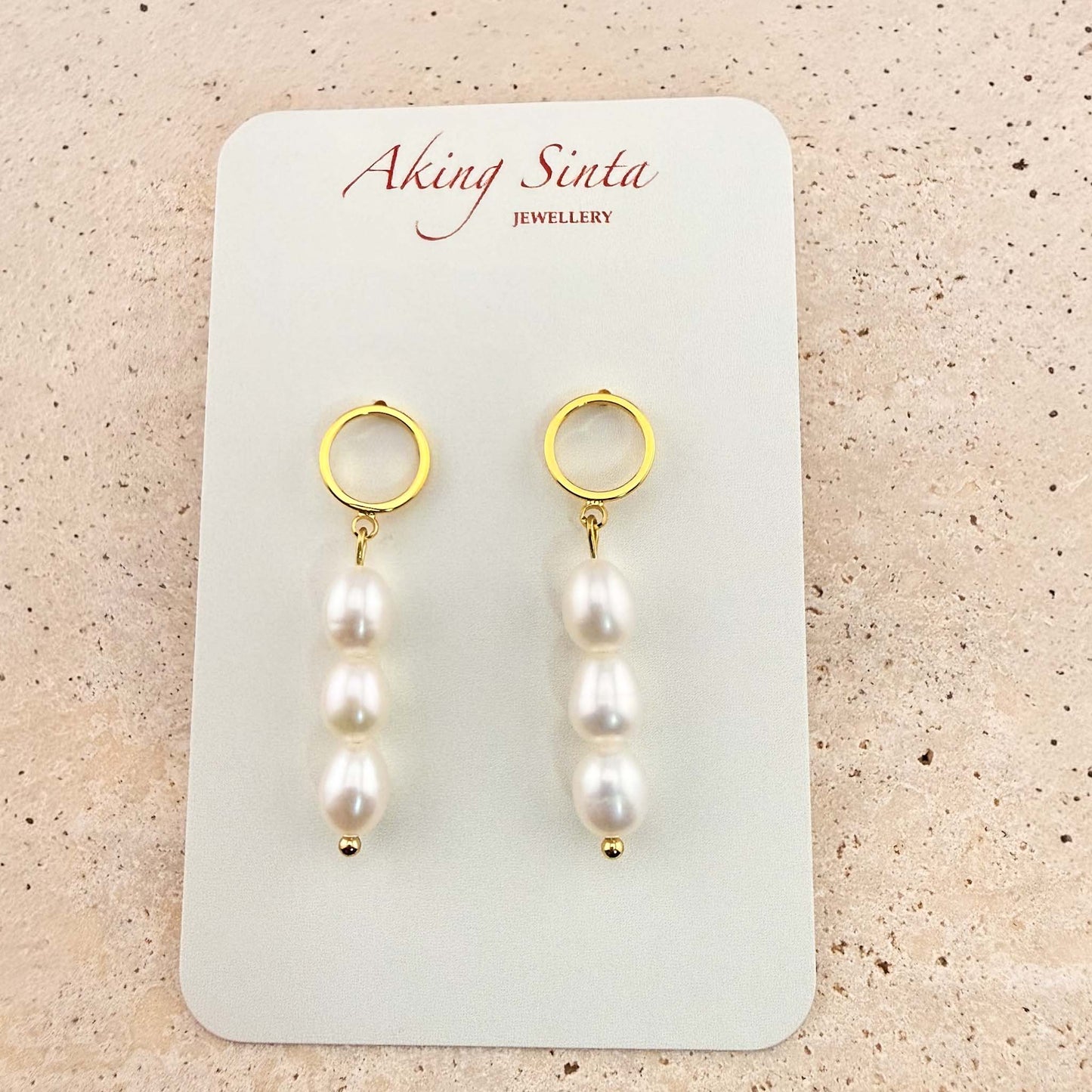 A pair of Aking Sinta triple pearl drop earrings on a white branded display card. Each earring features three white oval pearls hanging from a gold circle stud, set against a light stone background