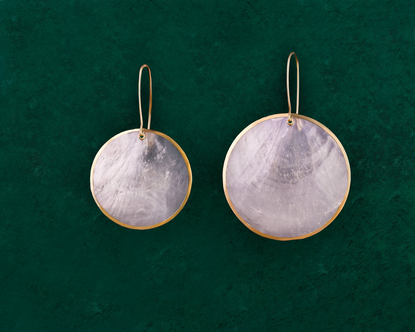 A pair of Aking Sinta Zoey’s Capiz Shell Wedding Earrings displayed on a dark green background; the large, round, iridescent white Capiz disks feature a subtle pearlescent glow and hang from polished gold hooks.