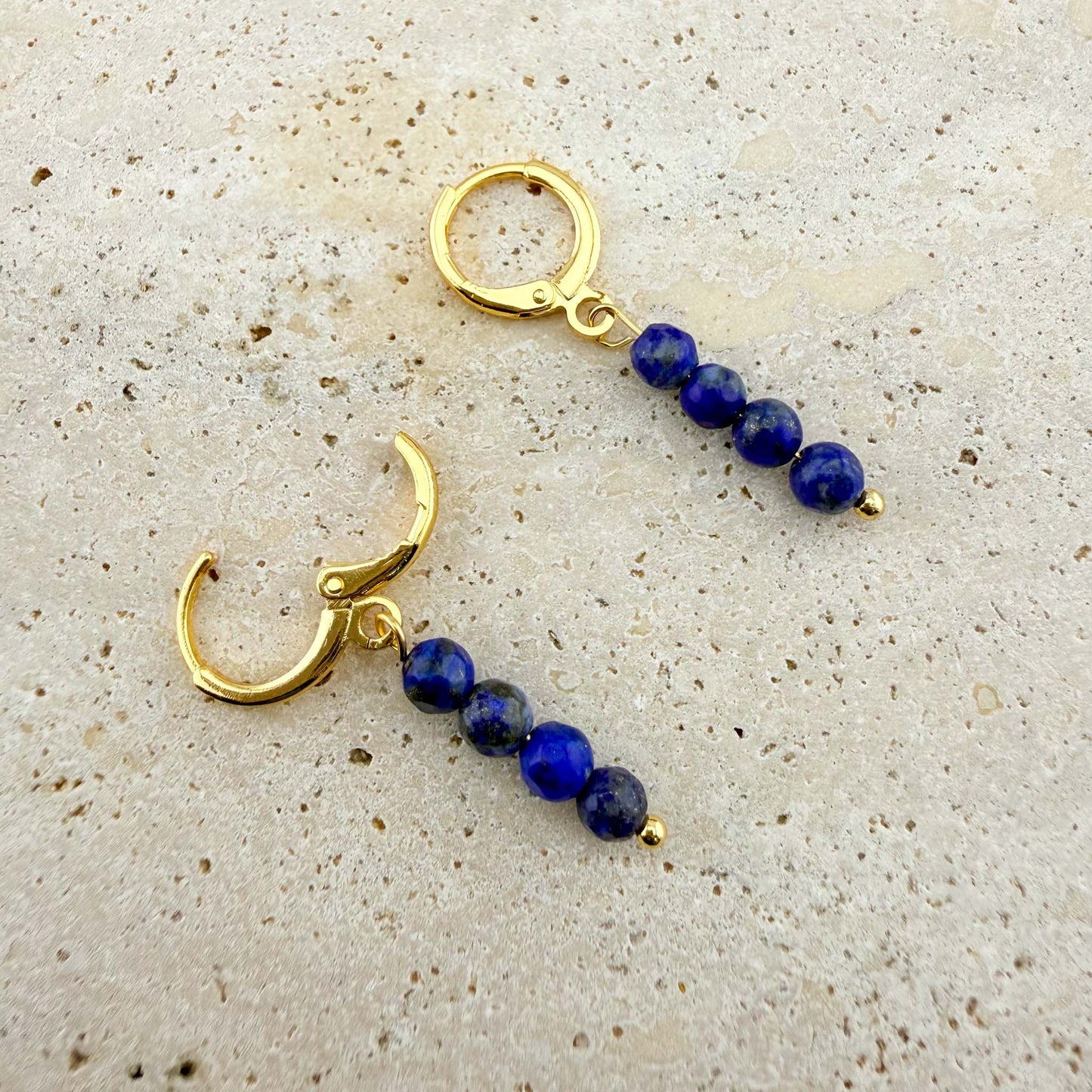 Macro view of gold Aking Sinta Lapis Lazuli Huggies on a white marble texture, featuring three stacked natural deep blue beads with one hinge clasp shown in the open position.