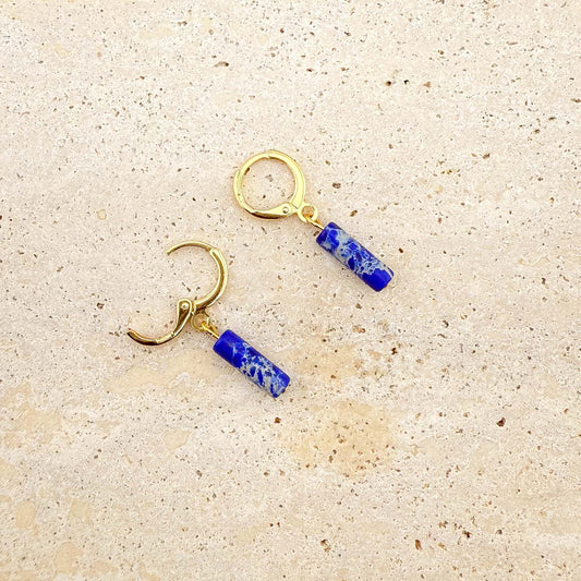 A detailed close-up of a pair of gold hoop earrings with cobalt blue cylinder charms resting on a light-colored speckled stone. The earring on the left has its loop clasped shut, while the earring on the right has its loop open at the bottom. The rich blue charms feature varied white and lighter blue natural stone patterns.