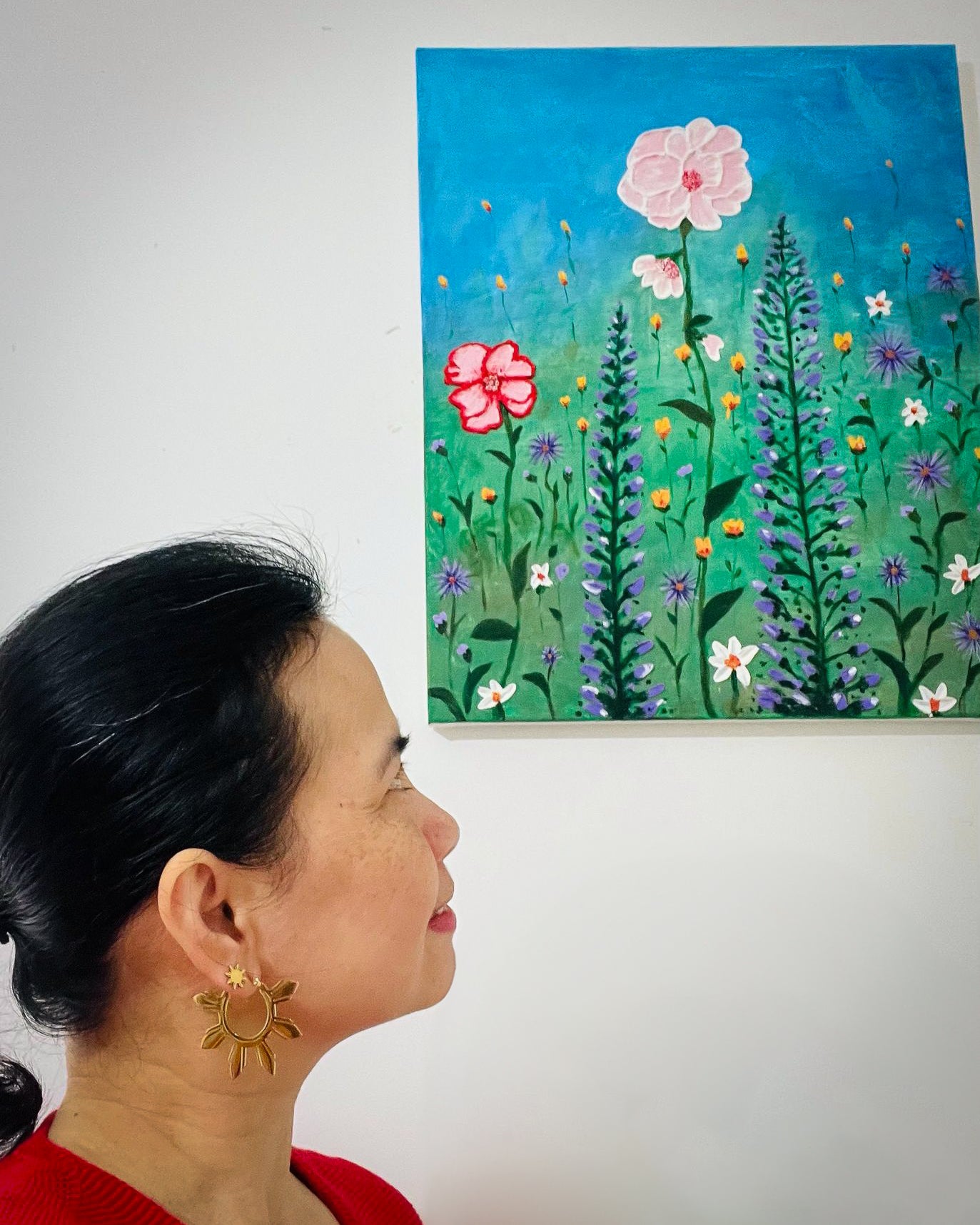 A woman in a red dress looking up at a flower painting while wearing Araw 8-ray sun stud earrings, capturing a sophisticated lifestyle moment of confidence and elegance.
