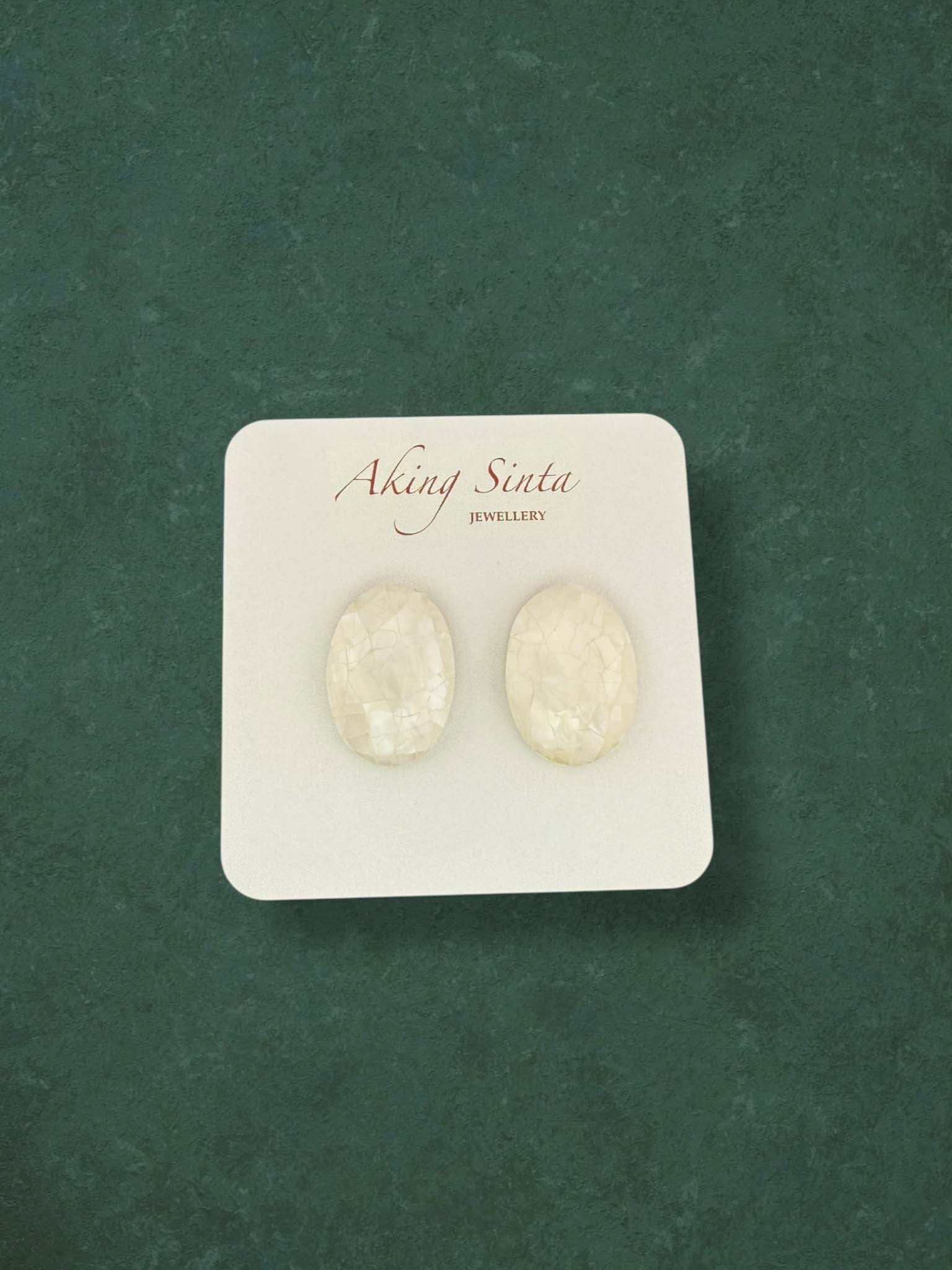 A pair of Aking Sinta white oval mosaic mother of pearl stud earrings. The handcrafted shell inlay design features a shimmering cracked-stone effect on a branded jewelry card