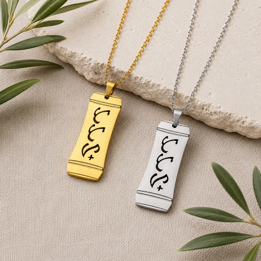 A product photograph showcasing two Aking Sinta vertical bamboo-style rectangular pendants in matte gold and silver finishes side by side. Both pendants feature the word "Tatay" (Father) intricately cut out in stylized traditional Baybayin script characters, displayed on a cream textured surface beside a light stone slab and olive leaves