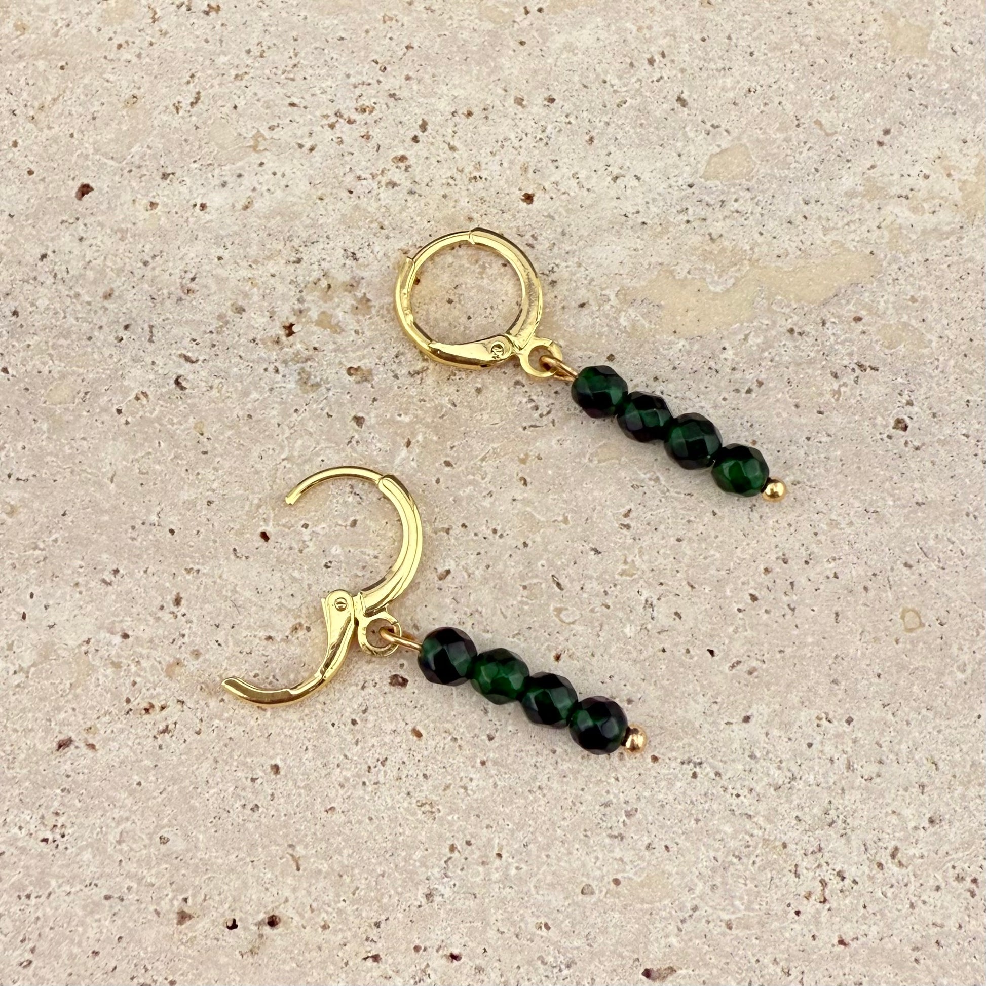 A pair of Aking Sinta gold huggie earrings with 4-bead Green Jade charms on a white textured stone; one gold clasp is open to show the easy-to-use hinge, while the other is closed, displaying the variegated green tones of the stacked beads.