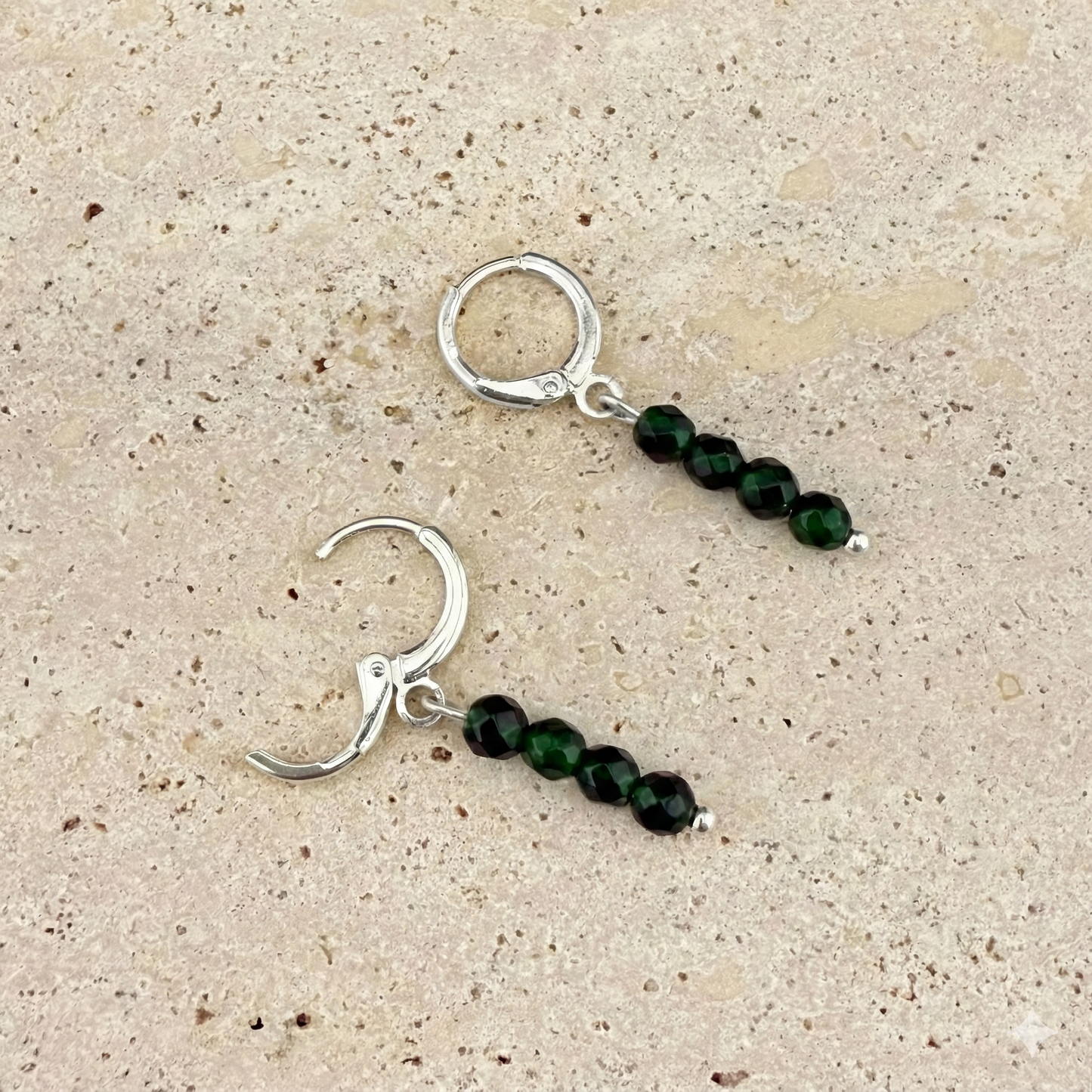 A pair of silver Aking Sinta huggie earrings on a white textured rock surface; each earring features a vertical strand of four round Green Jade beads with natural mossy patterns, suspended from a closed, polished silver hinged hoop.