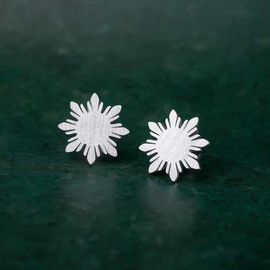 A pair of Aking Sinta Araw sterling silver stud earrings featuring the signature 8-ray sunburst design, displayed on a vibrant green background to highlight the cool-toned metallic shine