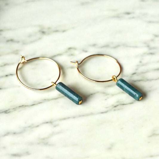 A pair of Aking Sinta gold hoop earrings on a white marble surface, featuring dangling vertical column-shaped African Jade stones in a deep, mottled forest green.