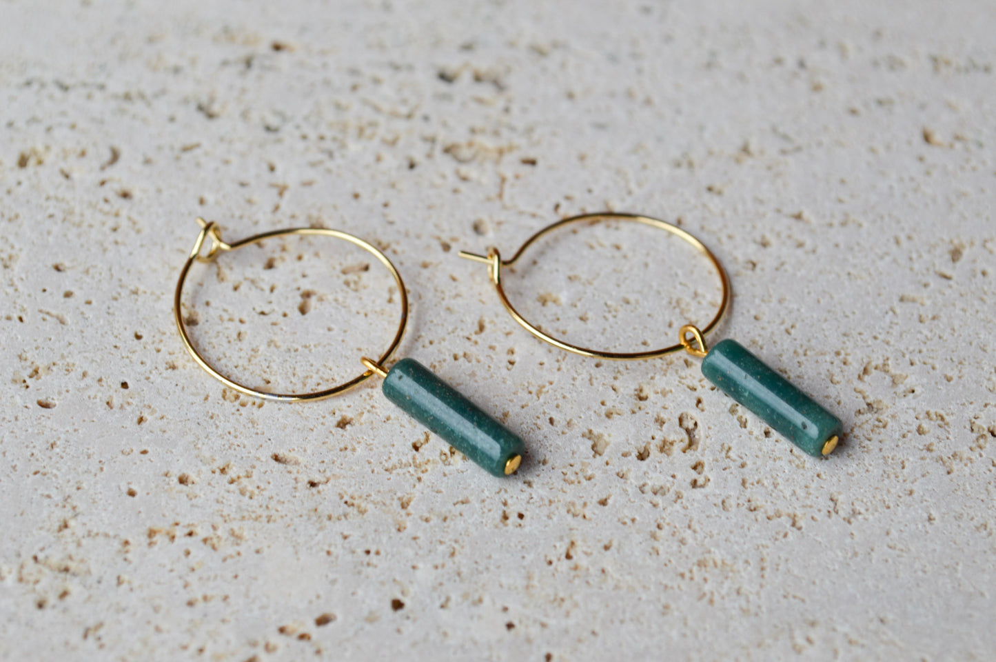A pair of gold hoop earrings with vertical African Jade column charms resting on a textured, porous white rock surface; the mottled forest green of the jade stands out against the rough, organic stone background.