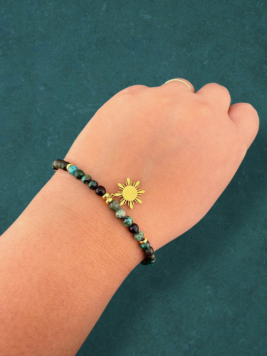 A close-up of an Aking Sinta African Turquoise beaded bracelet worn on a clenched fist; the earthy teal gemstones with dark black and brown mottling contrast with the polished gold 8-ray Araw charm and a gold extender chain with an engraved "Aking Sinta" tag.