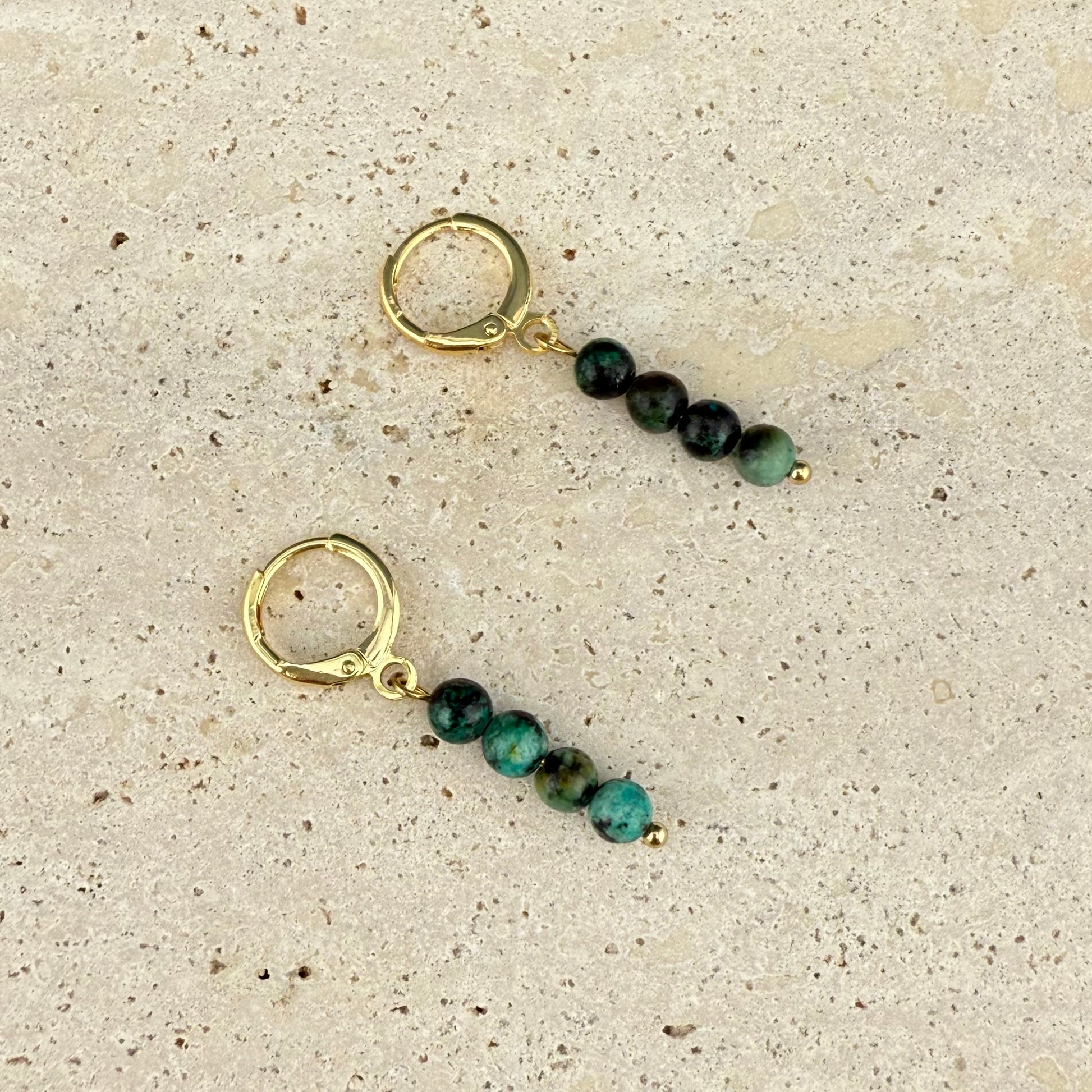 A pair of Aking Sinta African Turquoise gold huggie earrings with clasps closed, resting on a rough gray rock; the teal and black speckled beads are stacked vertically on the polished gold hoops.
