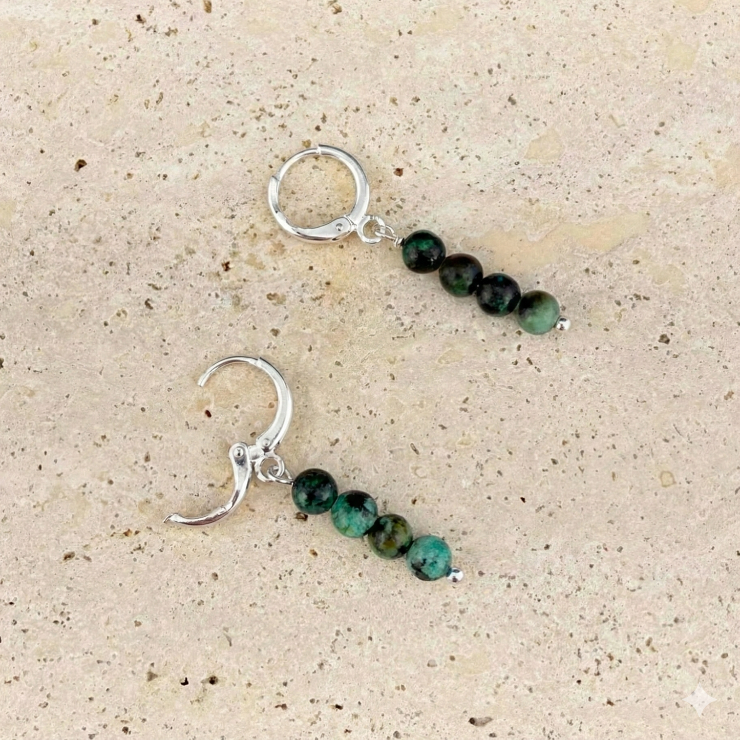 Silver variant of Aking Sinta African Turquoise huggie earrings on a textured gray rock; one silver hinge clasp is shown open, highlighting the vertical teal and black speckled beads.