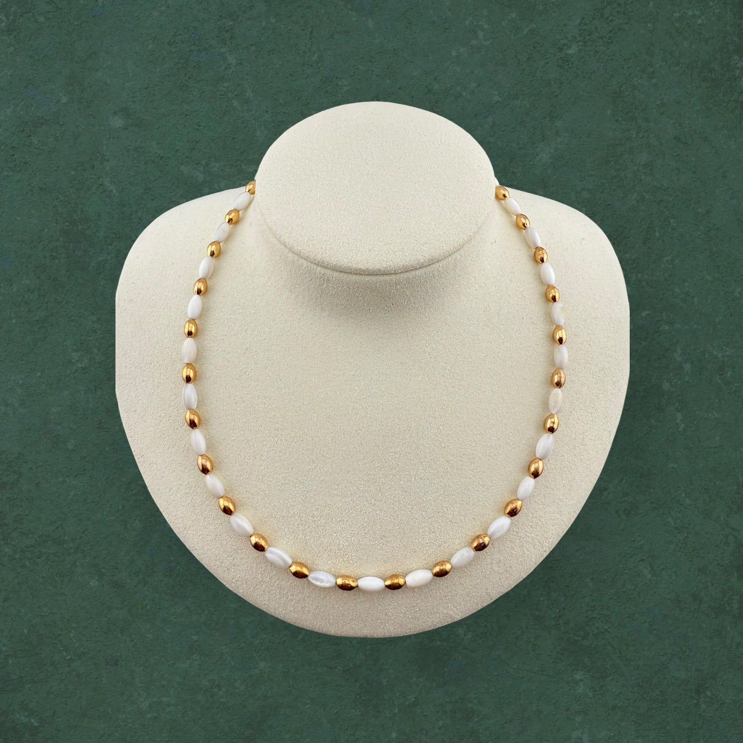 Aking Sinta rice bead necklace featuring an alternating pattern of white Mother of Pearl and gold beads, displayed on a jewelry bust against a dark green background.