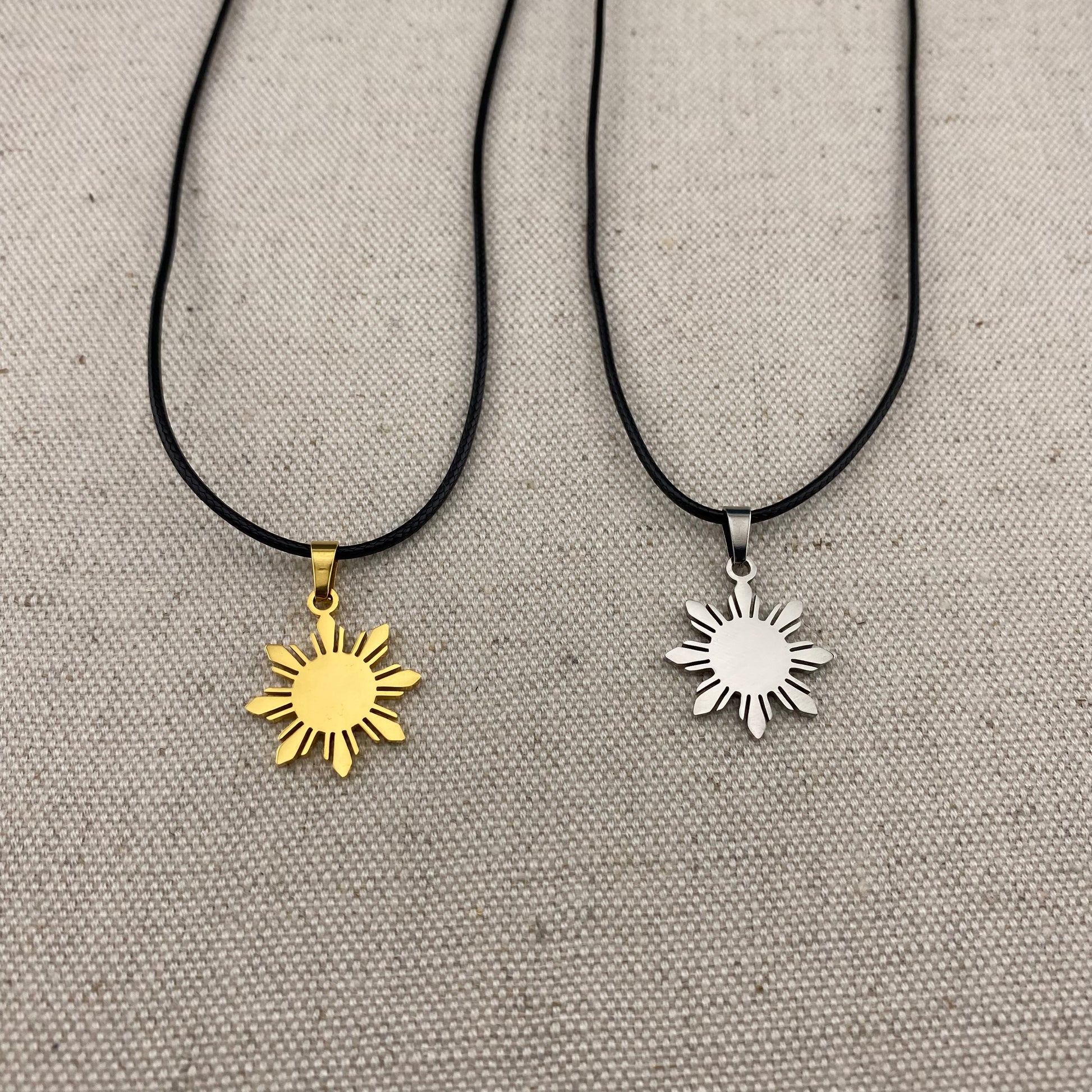 Aking Sinta Araw Cord Necklace with black cord and gold and silver Araw sun charms displayed on gray woven textured background, highlighting adjustable cord design and Philippine-inspired craftsmanship