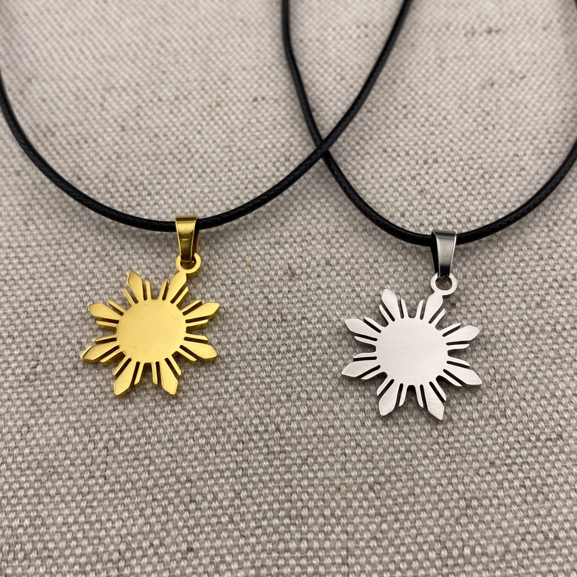 Aking Sinta Araw Cord Necklace with black cord in silver and gold variants displayed on gray woven textured background, highlighting adjustable cord design and Philippine-inspired sun charms