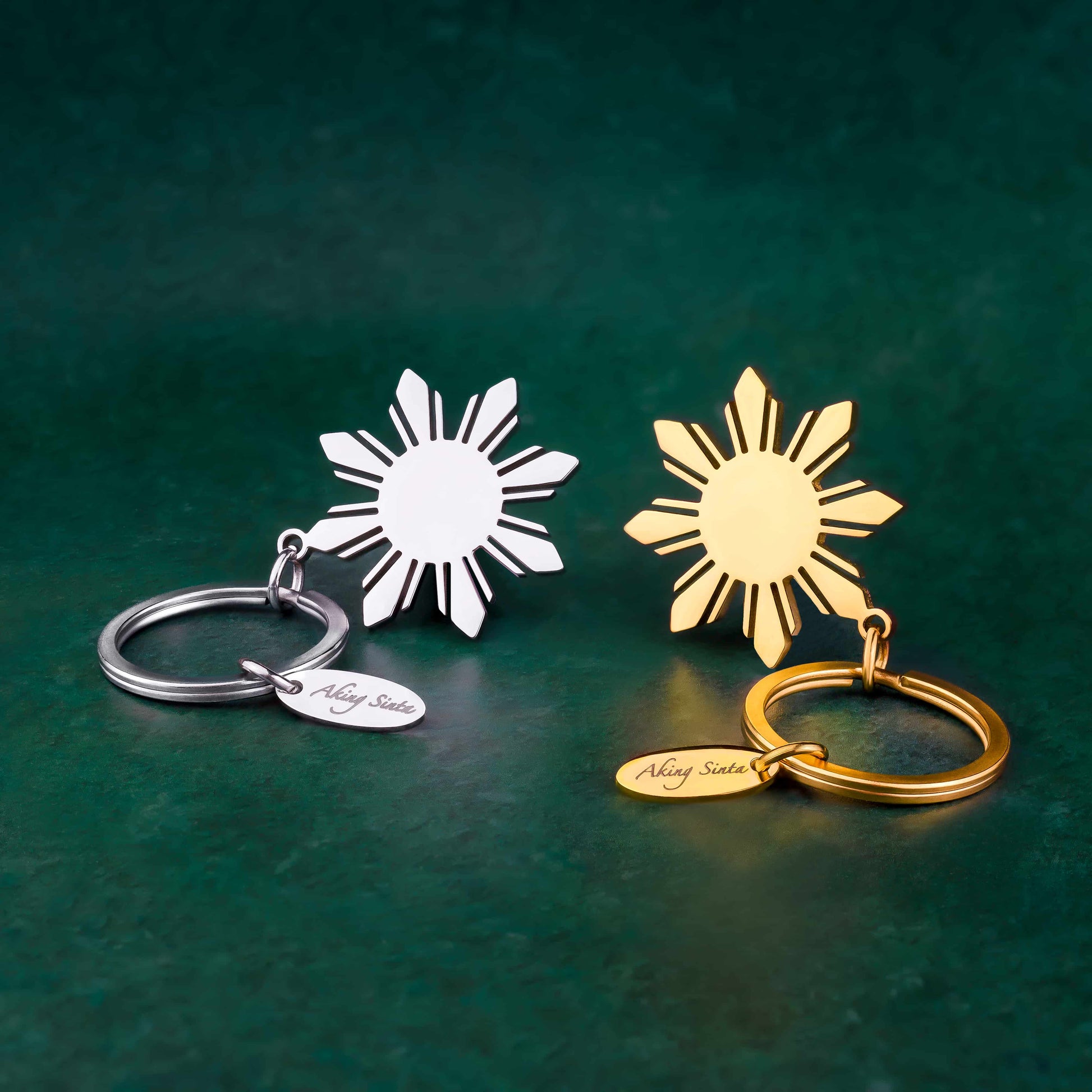 Both gold and silver variants of the Aking Sinta Araw key ring displayed on a lush green background; each featuring a large, polished 8-ray sunburst pendant attached to a heavy-duty metal ring