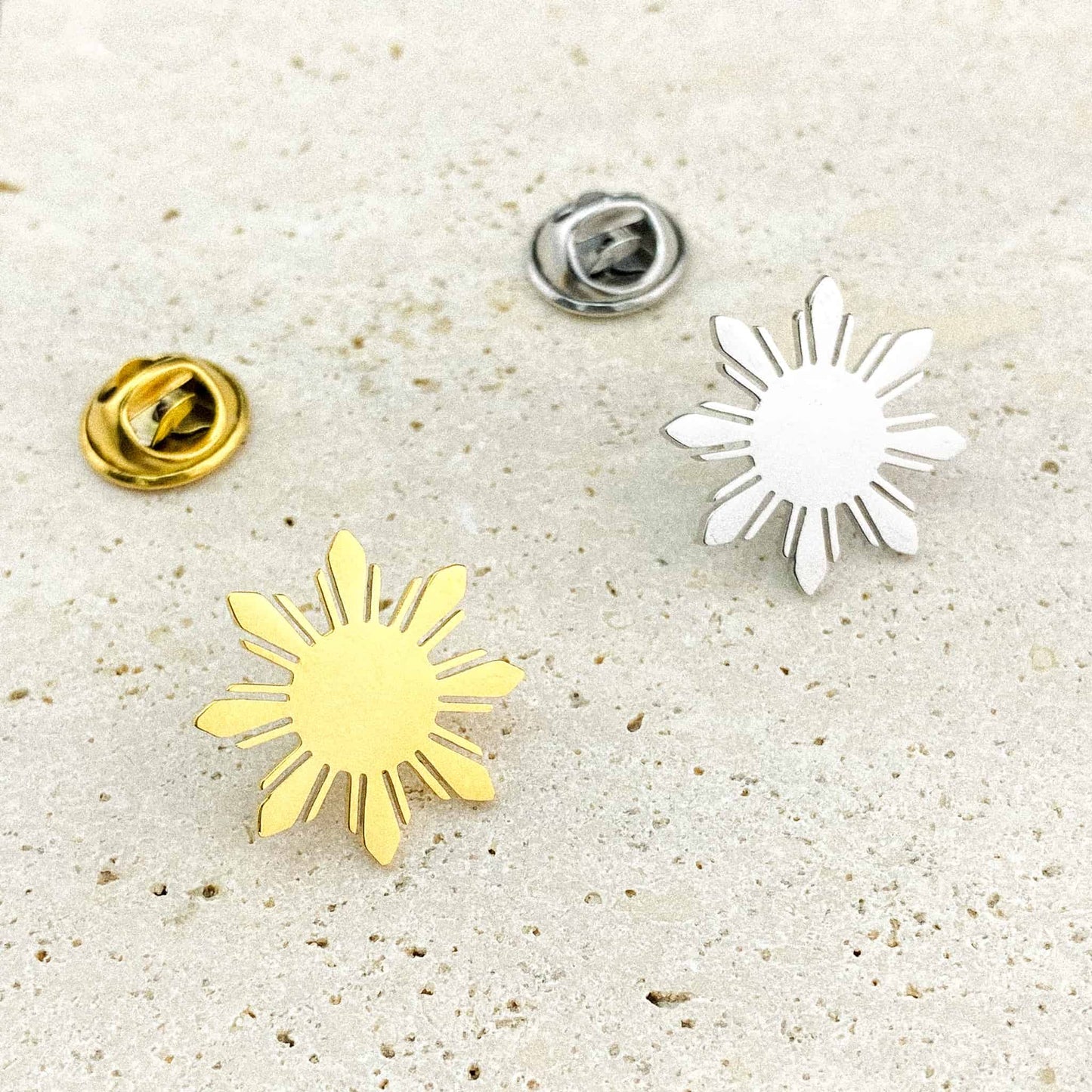 Two Aking Sinta Araw lapel pins in silver and gold variants placed on a white marble background, showcasing the intricate 8-ray sunburst design in a premium metallic finish.