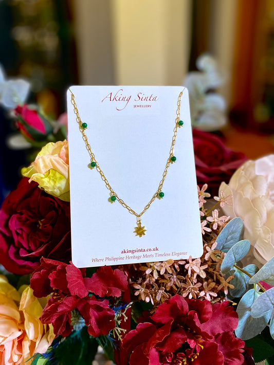 Close-up of an Aking Sinta paperclip necklace on branded cardboard; a dainty green jade bead is meticulously wired directly onto a gold chain link, positioned next to a hanging gold Araw sun charm, set against a backdrop of real flowers.