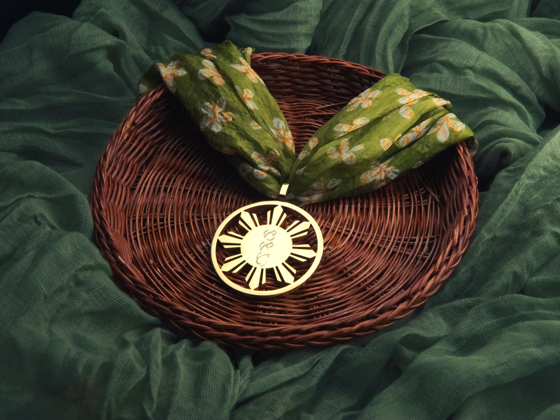 Aking Sinta gold Araw sun medallion displayed on a traditional woven rattan winnowing basket with green tropical flowers and a heavy gold chain.
