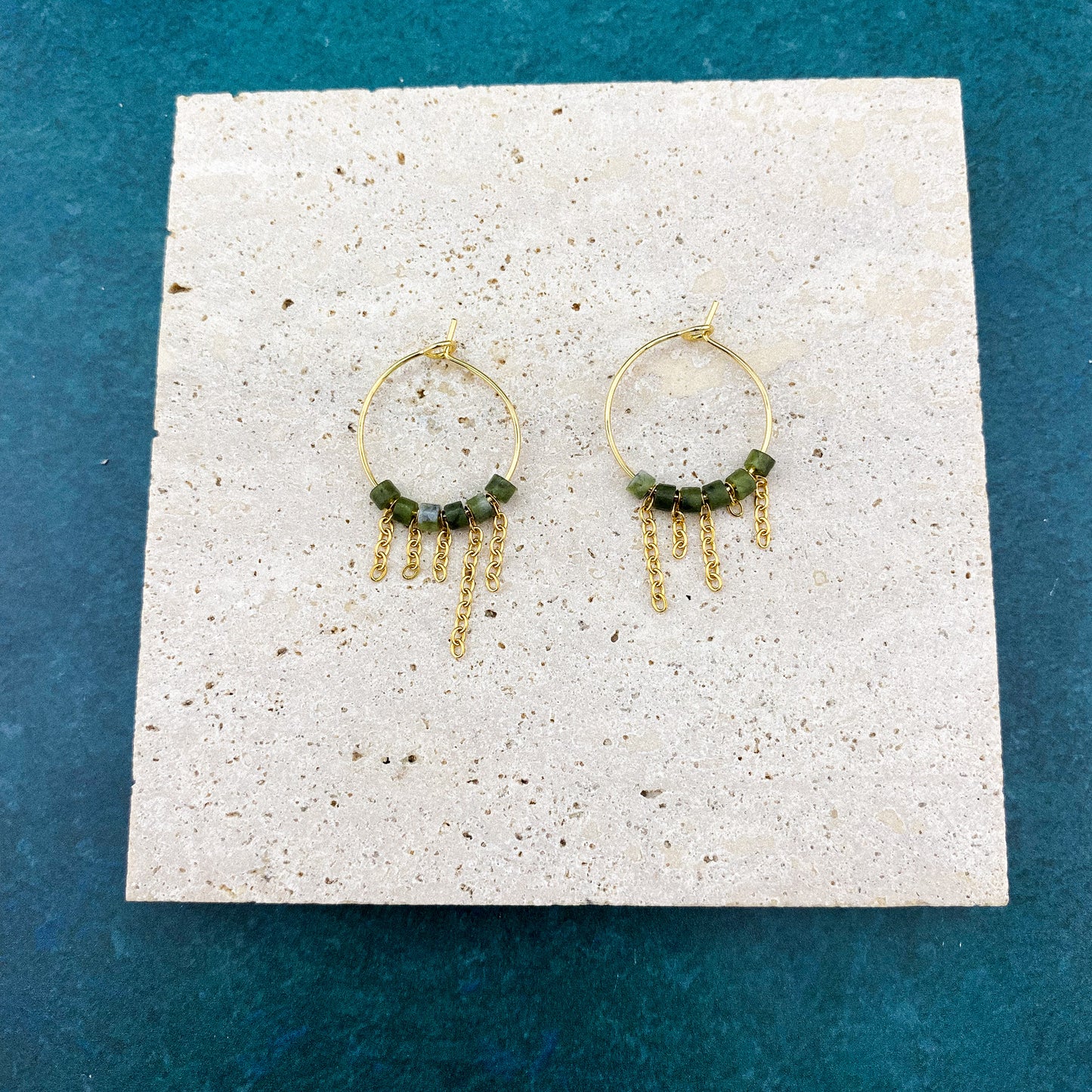 A pair of Aking Sinta gold hoop earrings with asymmetrical hanging fringes of Canadian Jade beads, laid flat on a smooth gray marble slab with white veining; the dark green stones are arranged in staggered, uneven lengths.