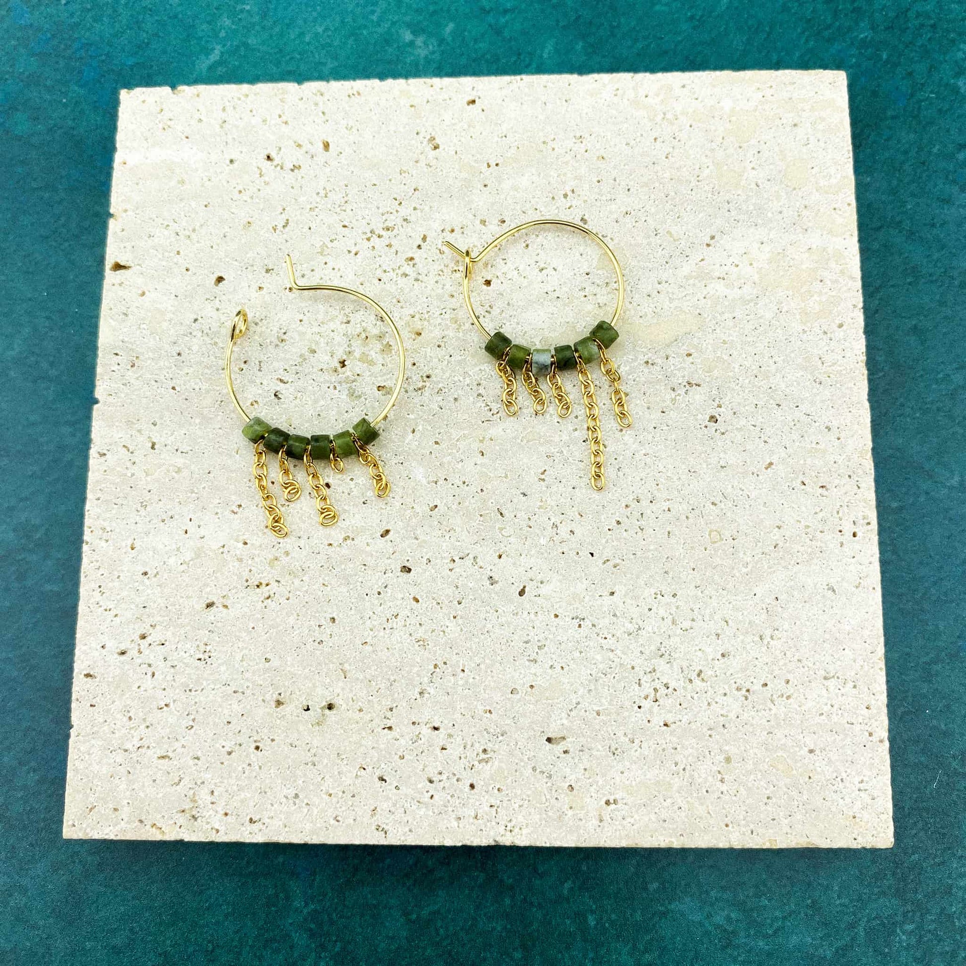 A pair of Aking Sinta gold hoop earrings with asymmetrical hanging fringes of Canadian Jade beads, laid flat on a smooth gray marble slab with white veining; the dark green stones are arranged in staggered, uneven lengths.