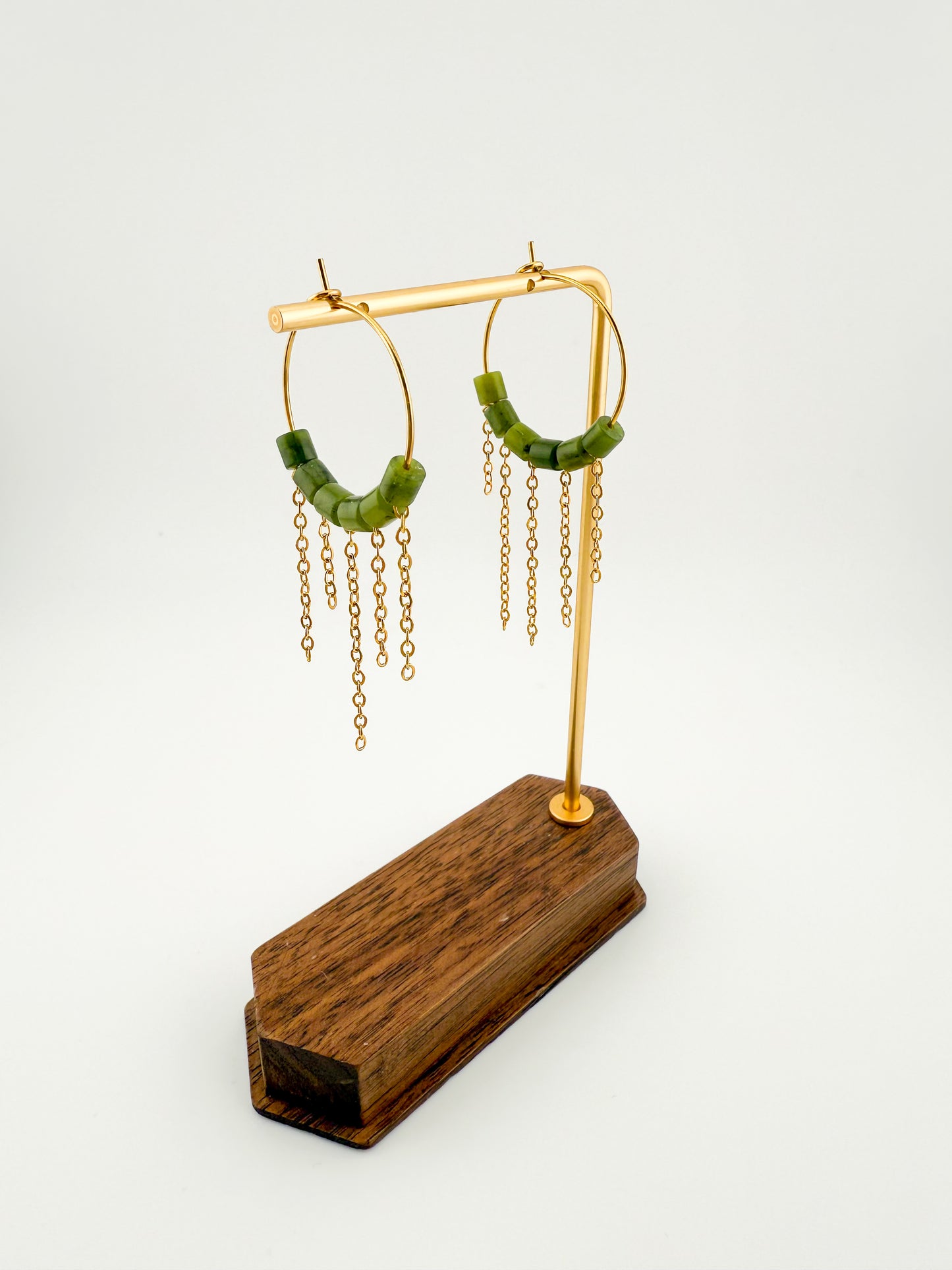 A pair of Aking Sinta gold hoop earrings with asymmetrical fringes of dark green Canadian Jade beads, displayed on a minimalist light wood jewelry stand; the beads hang at varying lengths for a modern, organic look.