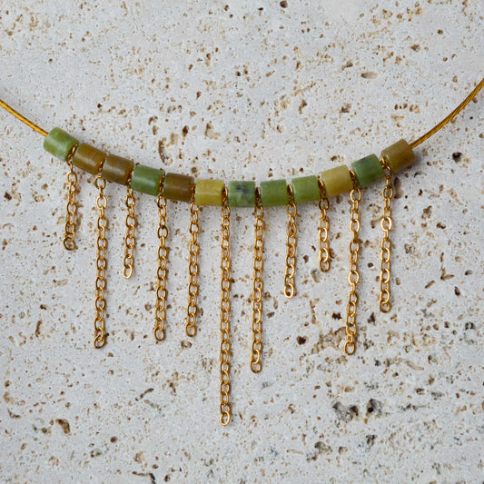 An Aking Sinta gold choker necklace with an asymmetrical fringe of varying lengths, laid flat on a gray marble slab; the design features a modern, uneven drop that creates a dynamic, architectural look.