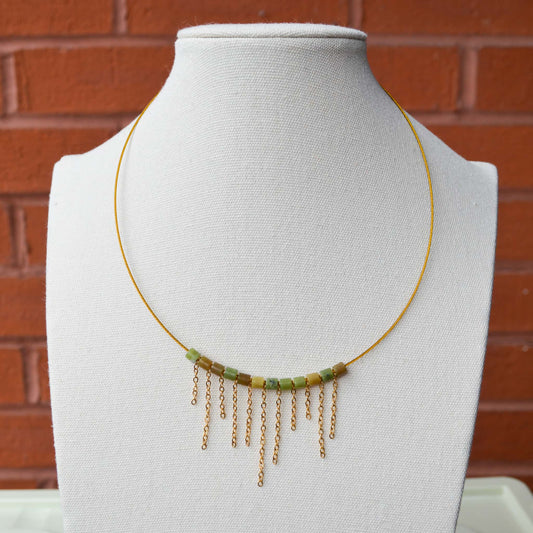 A gold Aking Sinta asymmetrical fringe choker displayed on a white neck bust; the necklace features gold chain drops of varying lengths that create a unique, uneven fringe across the front.