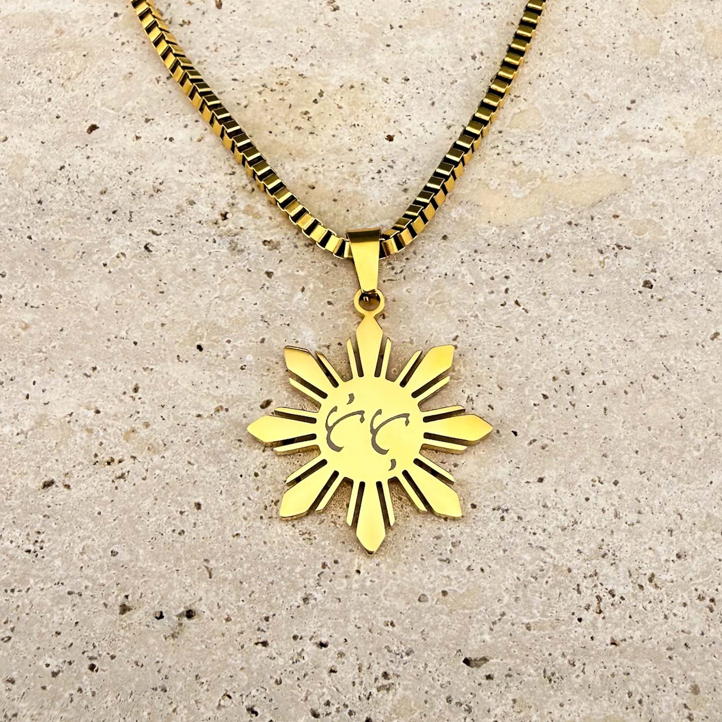 Close-up of a gold Aking Sinta Araw necklace on a marble background, featuring a fourth unique variant of bespoke Baybayin script engraving.