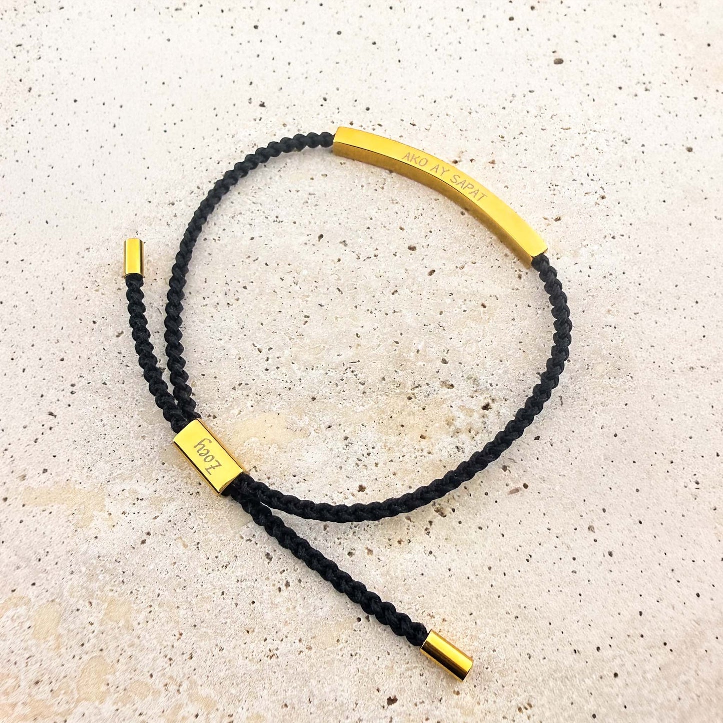 An Aking Sinta black woven cord friendship bracelet featuring a curved gold bar engraved with the Tagalog affirmation "AKO AY SAPAT" (I am enough). The adjustable bracelet includes gold-toned end caps and a custom-engraved slider with the name "Zoey," shown on a light stone surface