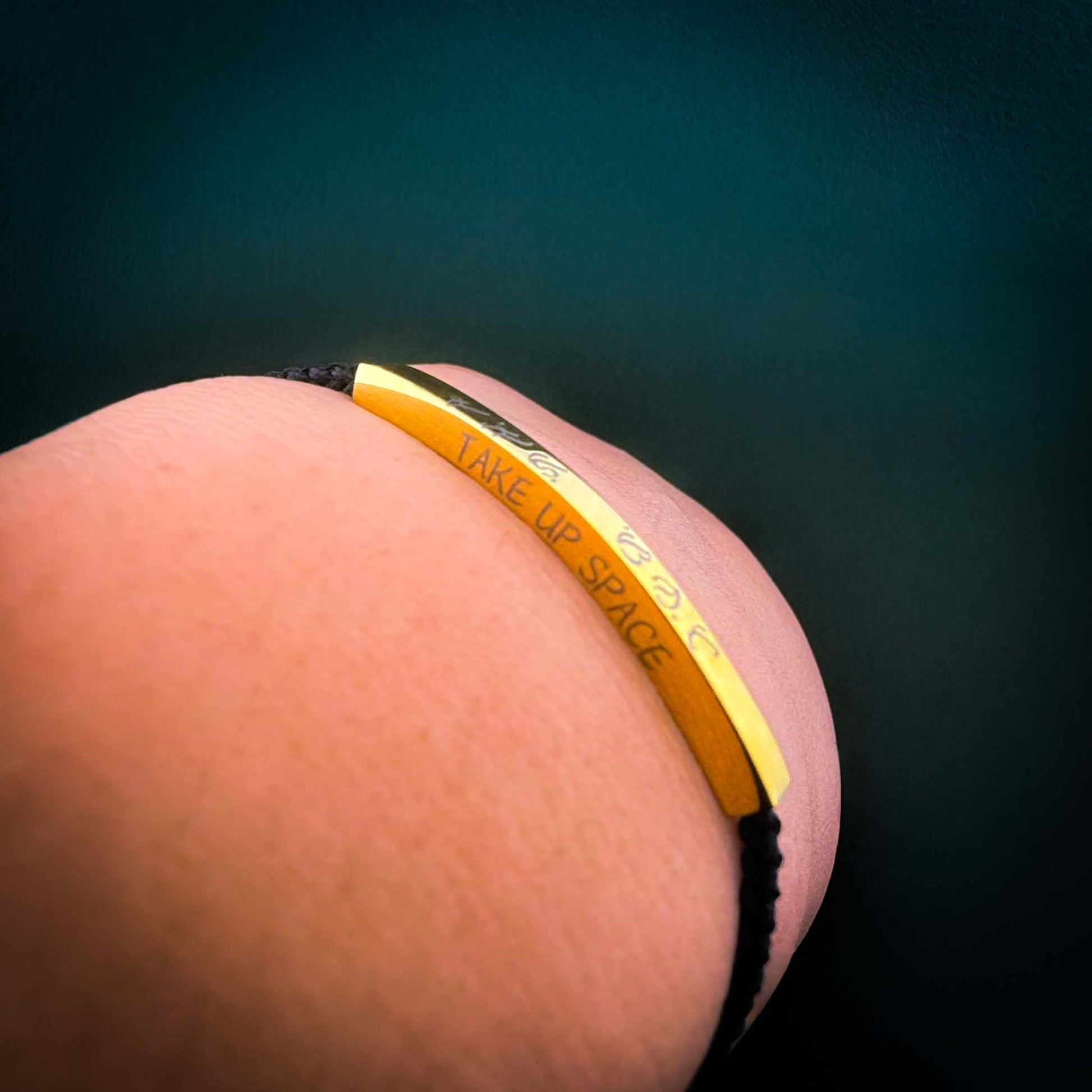 A close-up shot of an Aking Sinta black woven cord friendship bracelet being worn on a wrist. The curved gold bar is custom-engraved with the phrase "TAKE UP SPACE" alongside ancient Filipino Baybayin script, set against a dark teal background