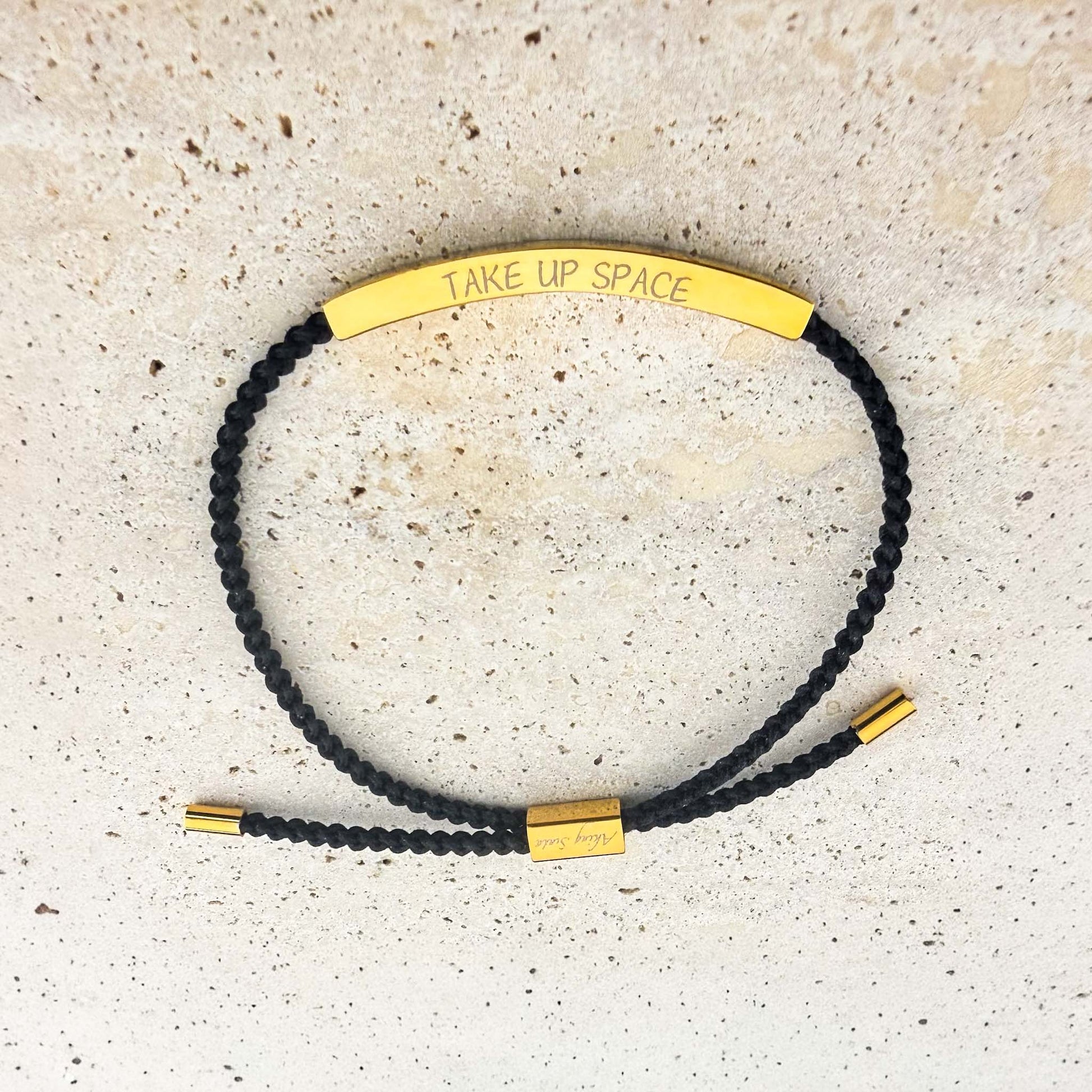 An Aking Sinta woven cord friendship bracelet in black featuring a curved gold bar. The bar is custom-engraved with the phrase "TAKE UP SPACE" in a minimalist font. The adjustable bracelet includes gold-toned accents and a branded slider, shown on a light-colored stone surface