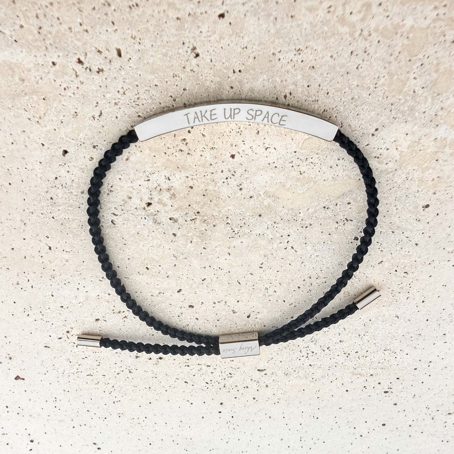 An Aking Sinta woven cord friendship bracelet in black featuring a curved silver bar. The bar is custom-engraved with the motivational phrase "TAKE UP SPACE." The adjustable design includes silver-toned end caps and a branded slider, shown on a light-colored natural stone surface