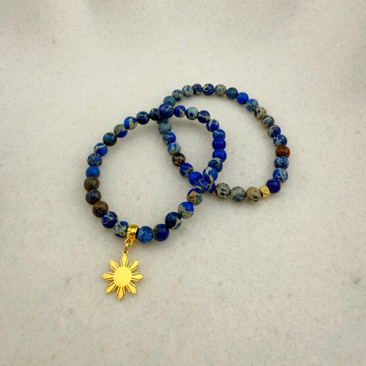 Two Aking Sinta blue Imperial Jasper stretch bracelets on a white background; both feature blue beads with white speckled patterns, one with a gold Araw charm and one without.