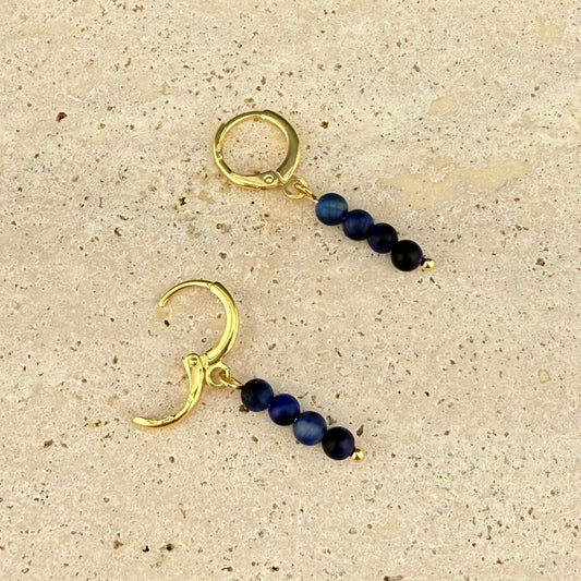 One Aking Sinta gold huggie earring shown with the hinge clasp open next to a closed one; the vertical stack of four Blue Tiger Eye beads and the tiny gold bottom bead are clearly visible on the marble surface.