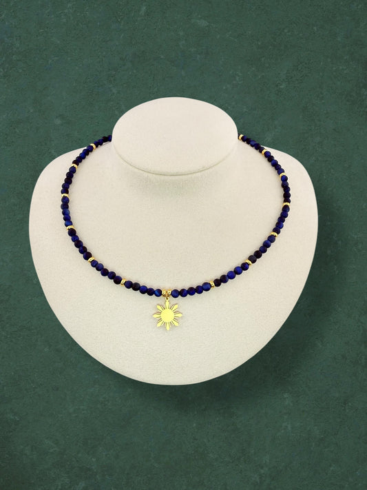Close-up of an Aking Sinta Blue Tiger Eye beaded necklace with a central gold 8-ray Araw sun charm; the dark blue stones show a cat-eye shimmer against a solid dark green background.