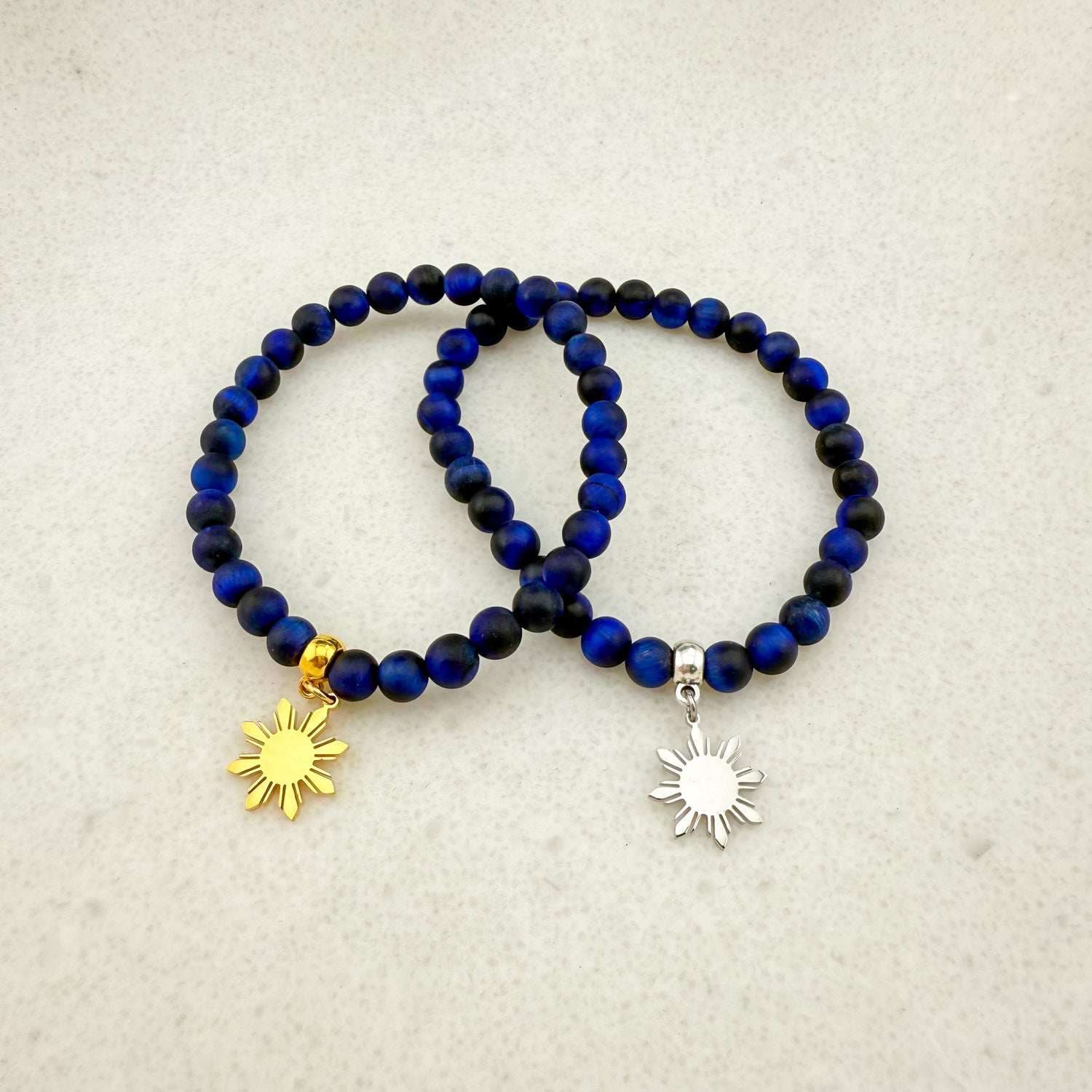 Two Aking Sinta Blue Tiger Eye stretch bracelets on a white background; featuring dark midnight-blue beads with a glossy finish, one with a gold Araw sun charm and one with a silver Araw sun charm.