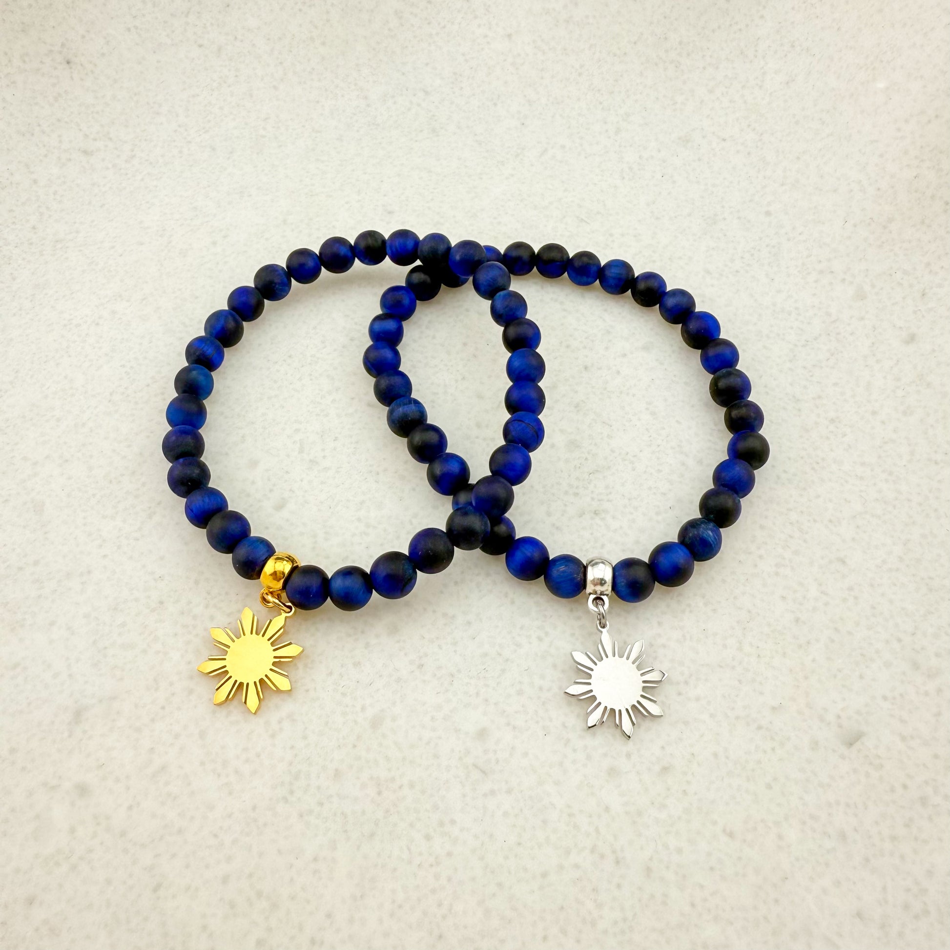 Two Aking Sinta Blue Tiger Eye stretch bracelets on a white background; featuring dark midnight-blue beads with a glossy finish, one with a gold Araw sun charm and one with a silver Araw sun charm.