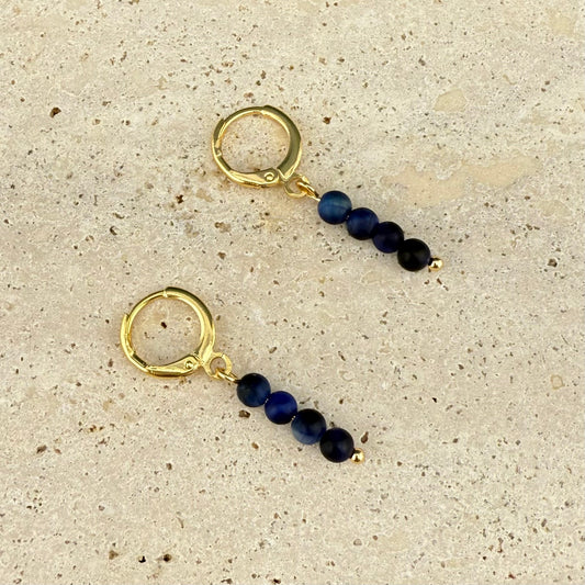 A pair of Aking Sinta gold huggie earrings featuring a vertical drop of four round Blue Tiger Eye beads; the stack of dark blue stones is finished with a tiny gold seed bead at the bottom, displayed on a white marble surface.
