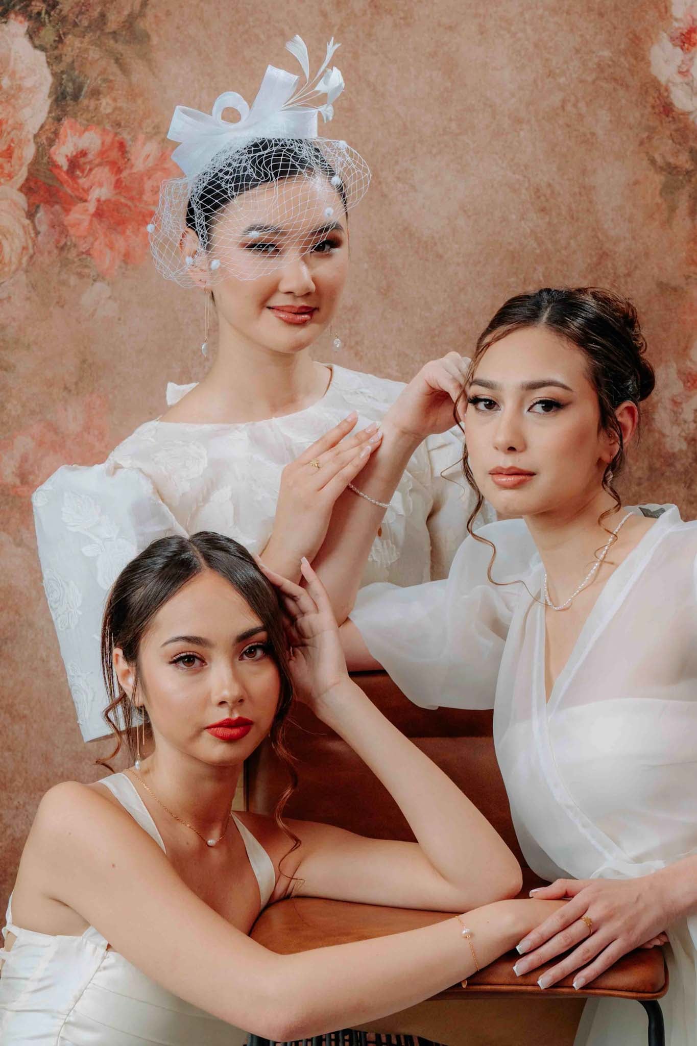 A sophisticated promotional photograph for the Aking Sinta jewelry collection, featuring three models styled in elegant white bridal-inspired attire. The models are adorned with delicate Aking Sinta gold chain threader earrings with single pearl drops, minimalist pearl necklaces, and fine gold chain bracelets. The scene is set against a warm, vintage-inspired floral background, showcasing the brand's timeless and romantic aesthetic
