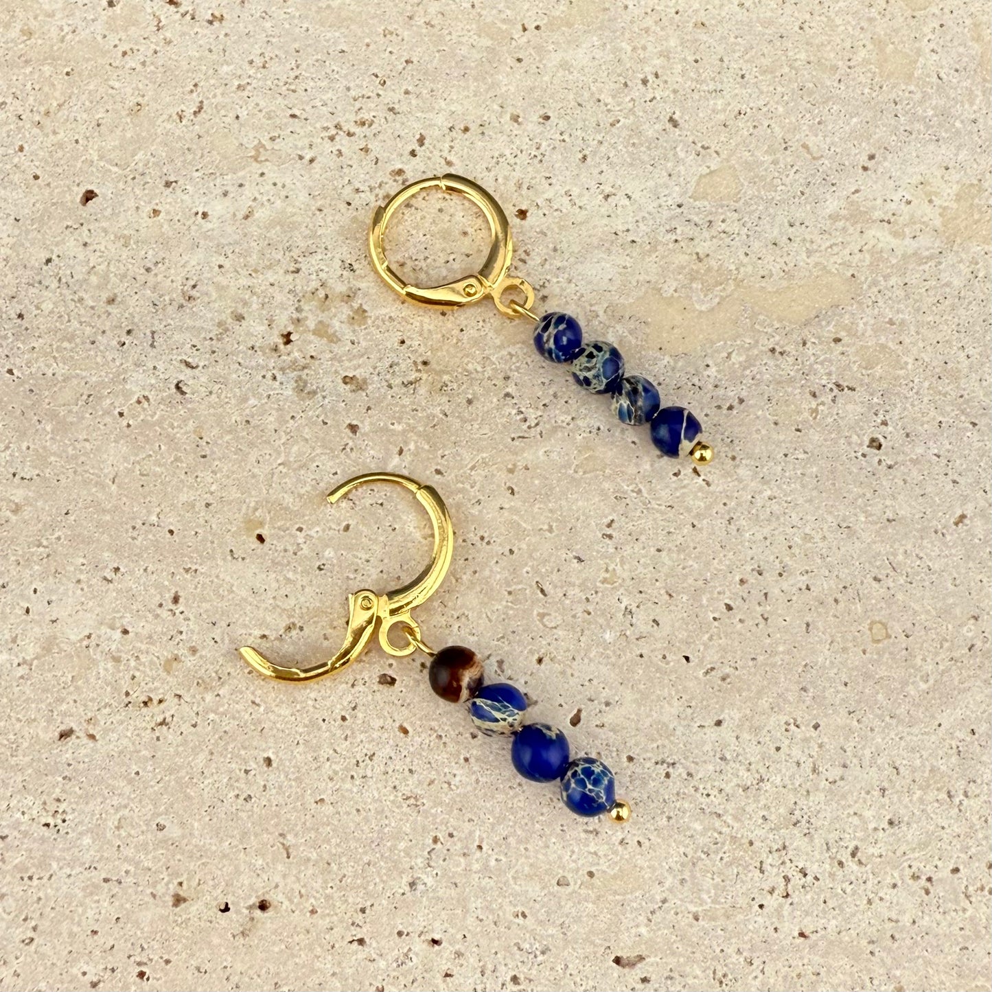 Gold variant of the Aking Sinta Brilliant Blue Imperial Jasper huggie earrings on a gray rock surface; one gold hinge clasp is open, showing the vibrant blue beads and the polished gold finish.