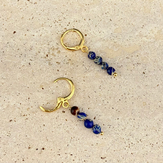 Gold variant of the Aking Sinta Brilliant Blue Imperial Jasper huggie earrings on a gray rock surface; one gold hinge clasp is open, showing the vibrant blue beads and the polished gold finish.