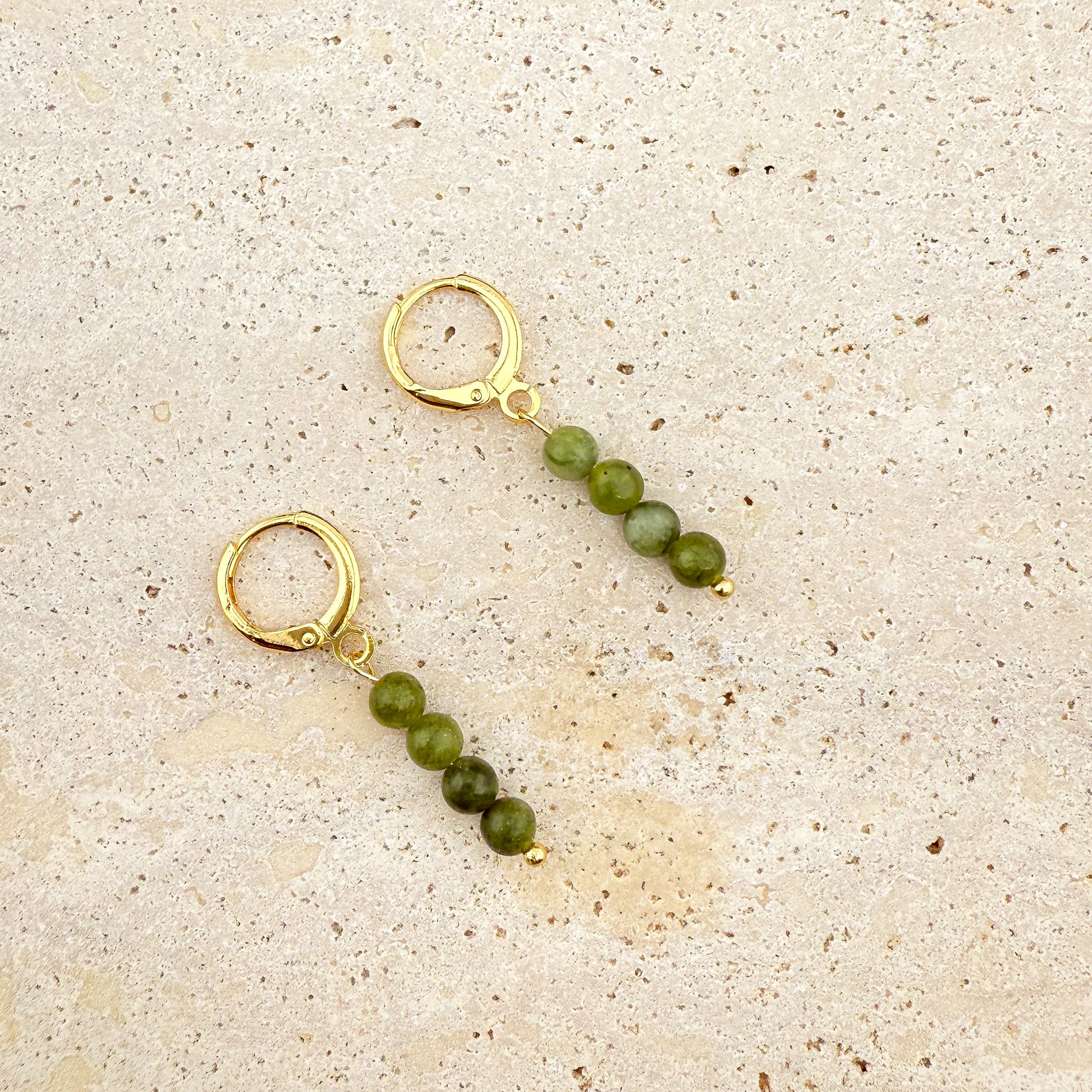 A pair of Aking Sinta gold huggie earrings on a white textured rock; each earring features a vertical drop of four round, polished Canadian Jade beads in a rich, dark translucent green.