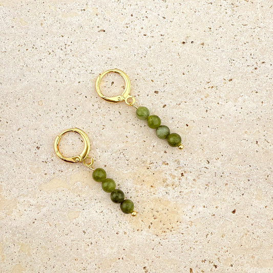 A pair of Aking Sinta gold huggie earrings on a white textured rock; each earring features a vertical drop of four round, polished Canadian Jade beads in a rich, dark translucent green.