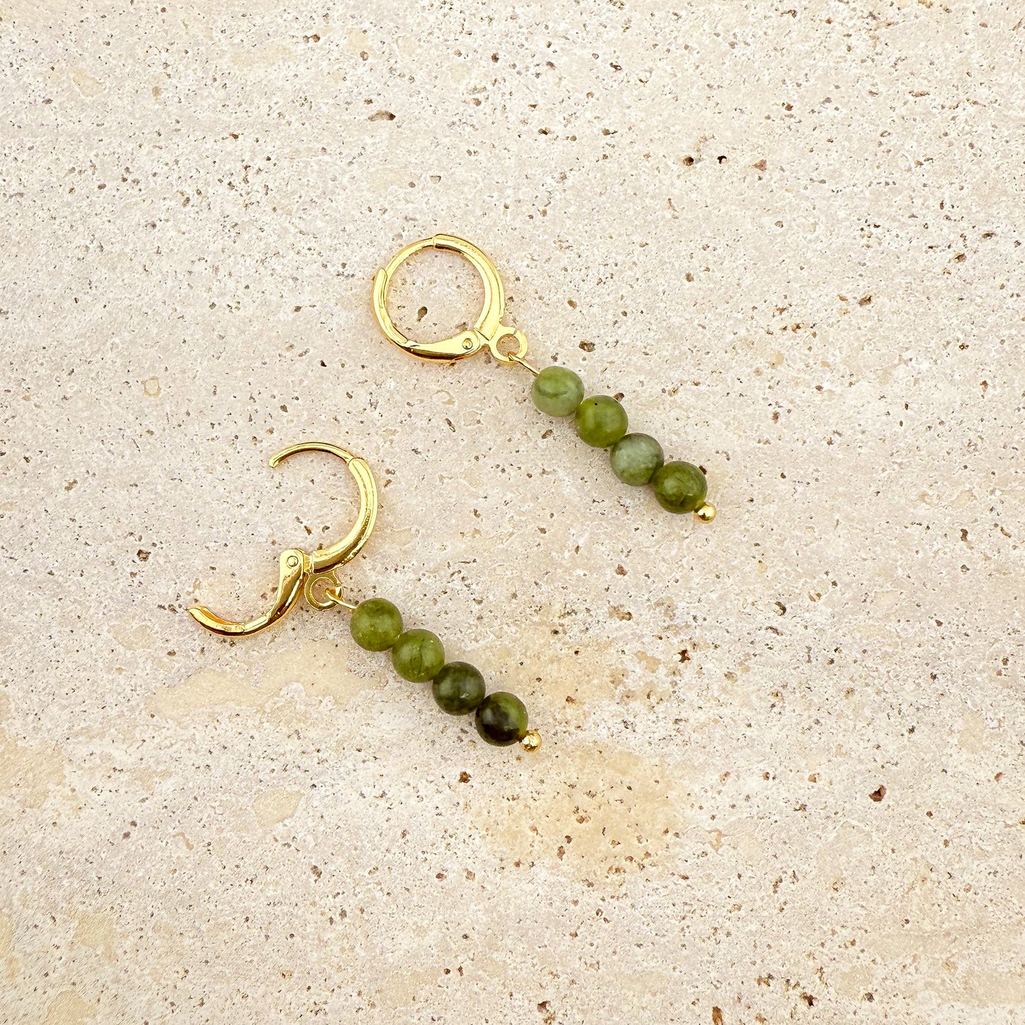 A pair of Aking Sinta gold huggie earrings with four-bead Canadian Jade charms on a white textured rock; one earring is shown with the gold hinge clasp open, while the other is closed, showcasing the long, dark green gemstone drop.