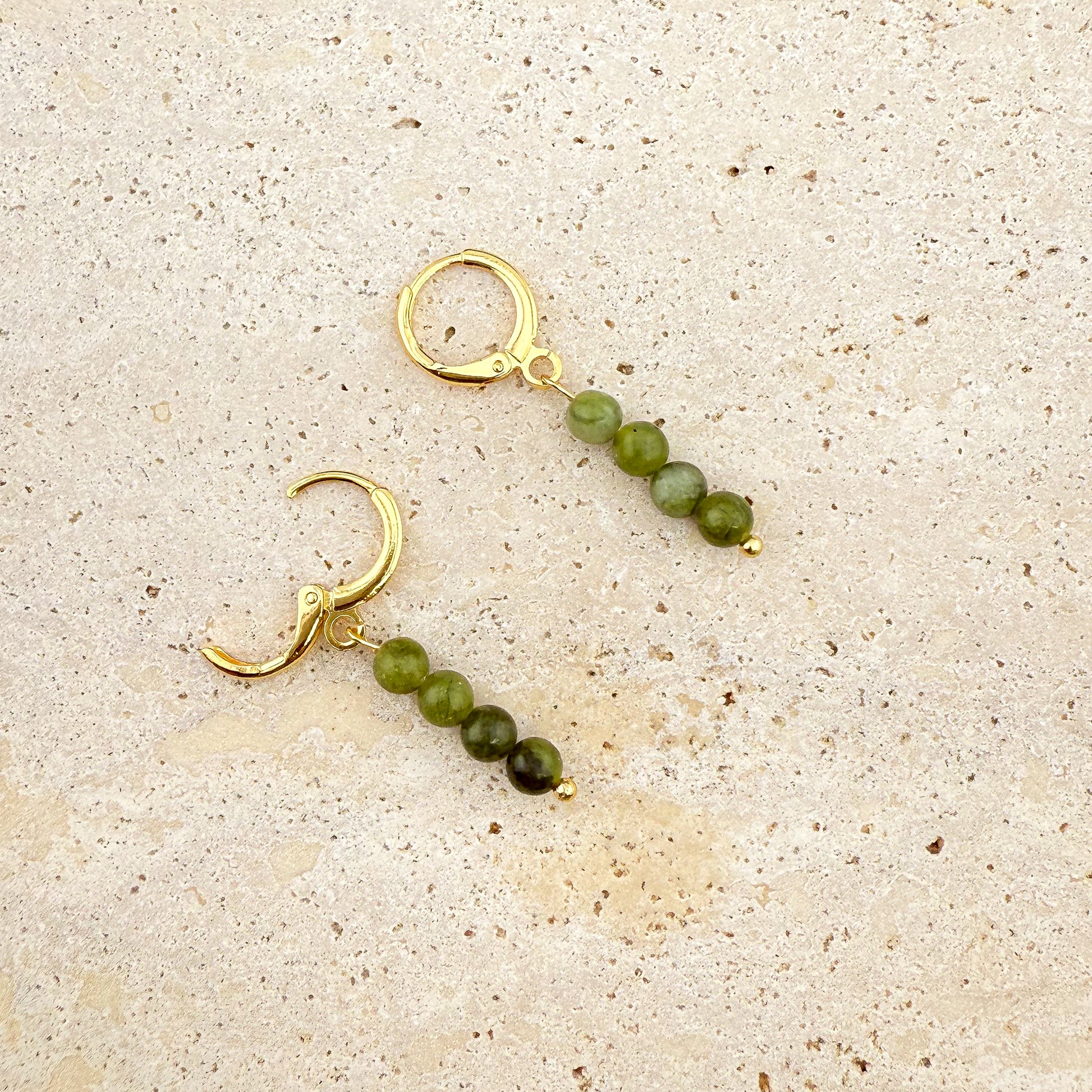 A pair of Aking Sinta gold huggie earrings with four-bead Canadian Jade charms on a white textured rock; one earring is shown with the gold hinge clasp open, while the other is closed, showcasing the long, dark green gemstone drop.